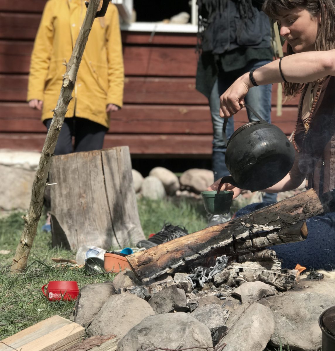 Ung kvinna i brun väst och blå byxor sitter vid lägereld i solen och häller upp kaffe som hon kokat över eld i en egengjord träkåsa