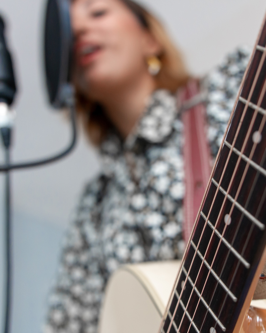 Ung kvinna i prickig blus sjunger i mikrofon och spelar gitarr