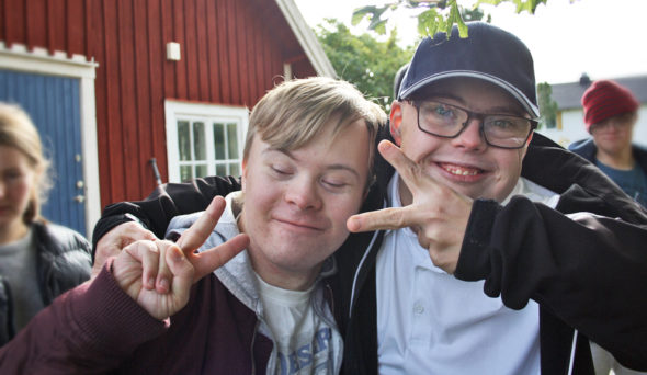 Två unga män med downs syndrom håller varandra om axlarna och ler in i kameran samtidigt som de håller upp två fingrar vardera i fredstecken