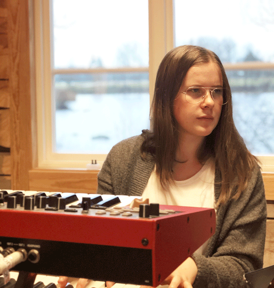 Ung kvinna spelar keyboard. i bakgrunden syns en sjö genom fönstret.