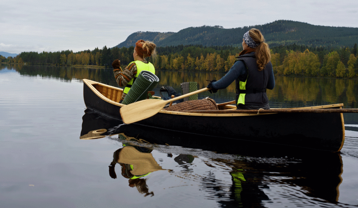 Två kvinnor paddlar kanot mot horisonten. Blank sjö i fjällmiljö