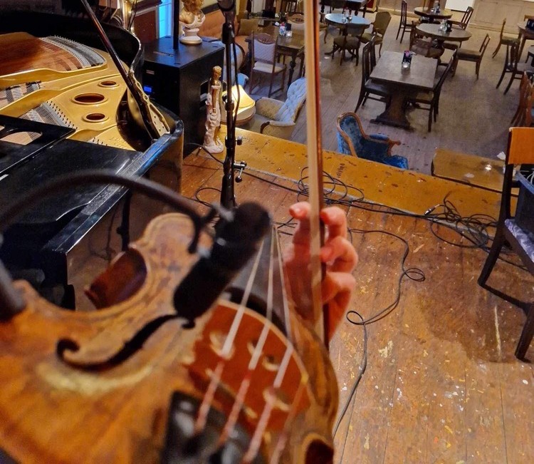 I förgrunden en fiol och en hand som håller i en stråke. I bakgrunden en scen och ett golv med stolar och bord där publik kan sitta.